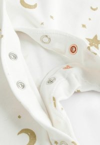Weißer Stoff mit goldenen Sternen und Halbmondmustern, mit metallischen Druckknöpfen in Silber und Roségold entlang der Öffnung.