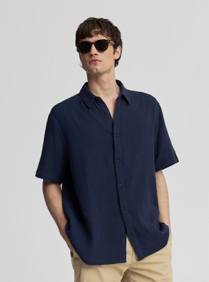 Giovane uomo che indossa occhiali da sole scuri, una camicia blu navy a maniche corte e bottoni, e pantaloni beige, in piedi con le mani nelle tasche.