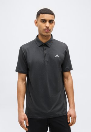 Rövid hajú fiatal férfi sötétszürke Adidas galléros pólóban, egyszínű világos háttér előtt, előre nézve áll.