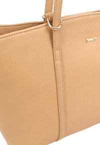 Sac fourre-tout en cuir beige avec une finition texturée, mettant en avant des accents cousus et une étiquette en métal. Comprend des poignées doubles pour le transport.