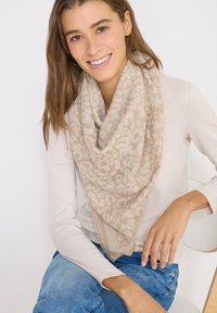 Femme souriante aux longs cheveux bruns portant une écharpe à motifs beige, un haut à manches longues clair et un jean bleu, assise avec un bras posé sur une chaise.