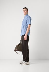 Lichtblauw kortemouwen overhemd met knopen, zwarte broek en witte sneakers. Houdt een bruine duffelbag met donkere banden vast.