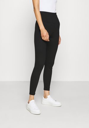 Woman wearing black high-waist leggings and white sneakers, standing against a plain white background.