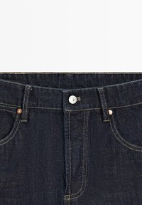 Dark blue denim jeans waistband with metal button and visible yellow stitching around front pockets and belt loops.