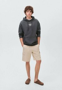 Felpa con cappuccio grigio scuro con tasca anteriore e testo, abbinata a pantaloni corti beige chiaro con tasche laterali. Scarpe marroni senza lacci.
