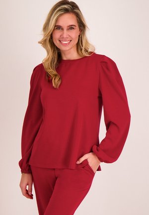 Lachende vrouw met blond haar draagt een rood shirt met lange mouwen en opgeblazen schouders en bijpassende rode broek, staand met één hand in haar zak.