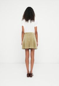 Donna con capelli neri ricci in piedi di fronte a un muro bianco, indossa una maglietta bianca, una gonna in velluto a coste beige con cerniera sul retro e scarpe in pelle marrone.