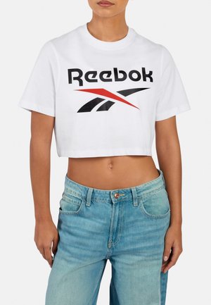 Vrouw draagt een wit cropped Reebok T-shirt en lichtblauwe high-waisted jeans, staand met ontspannen armen langs haar zij.