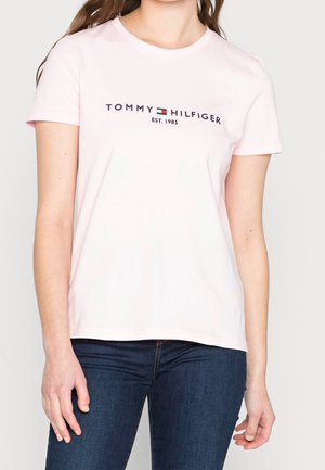 Kobieta w jasnoróżowej koszulce Tommy Hilfiger z logo i ciemnoniebieskich jeansach, stojąca na jednolitym tle.
