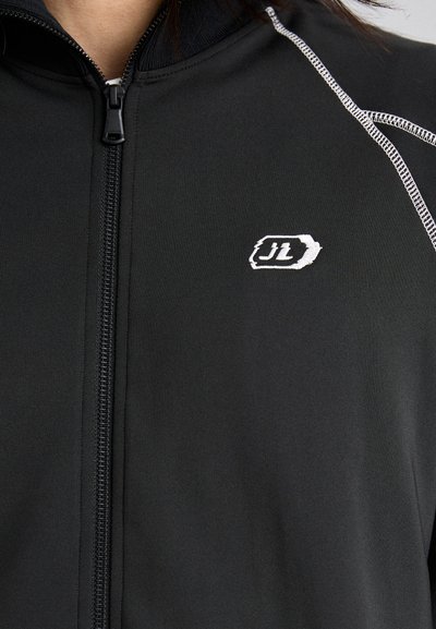 Juodas užtrauktuku užsegamas švarkas, pagamintas iš lygaus audinio su aukštu apykakle. Yra baltos siuvinėjimo akcentai ir mažas išsiuvinėtas logotipas krūtinėje.