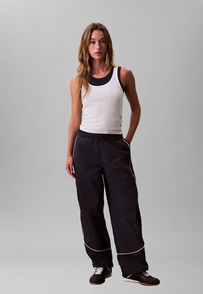 Jeune femme debout, une main dans la poche, portant un débardeur blanc par-dessus une chemise sans manches noire, un pantalon de sport noir avec une bande blanche, et des baskets noires.