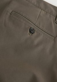 Barna nadrág hátsó zsebe egy fekete gombbal és vízszintes hasítékzseb nyílással.