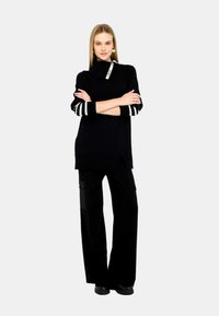 Maglione oversize nero con polsini a strisce bianche e uno spacco laterale, abbinato a pantaloni cargo neri larghi con tasche laterali. Scarpe nere.