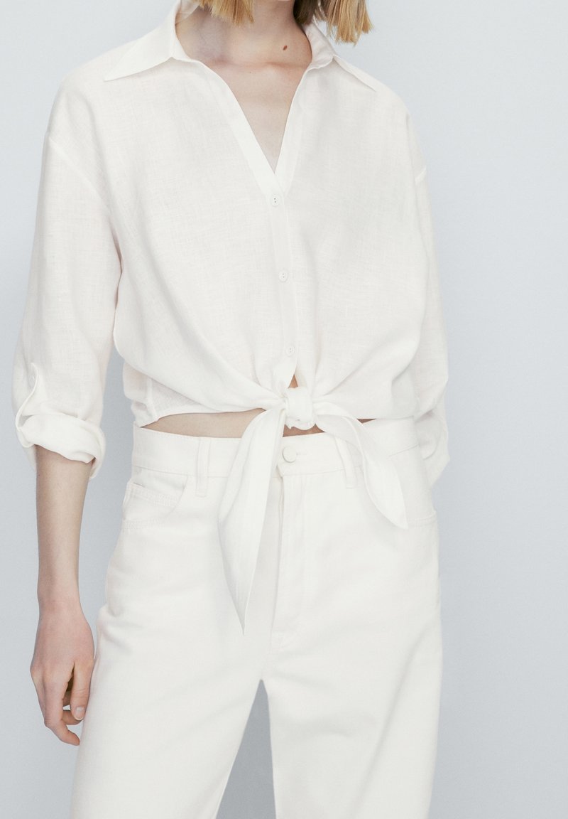 Witte, gestructureerde linnen blouse met een V-hals en knoopsluiting, geknoopt in de taille, gecombineerd met witte hoge taille broek.