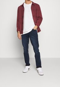 Röd skjorta med knappar över en vit t-shirt med texten "OPTIMISM", tillsammans med mörkblå jeans och vita sneakers med svarta detaljer.