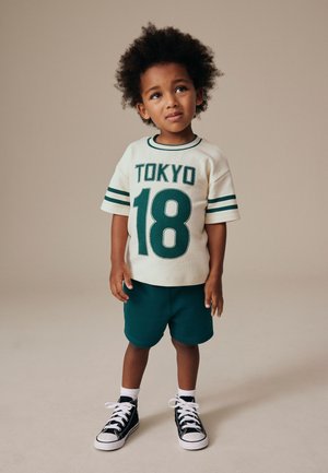 Młody chłopiec z kręconymi włosami, ubrany w biały t-shirt "Tokyo 18", zielone spodenki, białe skarpety i czarne wysokie trampki, stoi na beżowym tle.