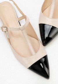 Zapatos slingback de cuero beige con punta afilada de charol negro brillante, que destacan por una correa en el tobillo con hebilla y detalles de costura visibles.