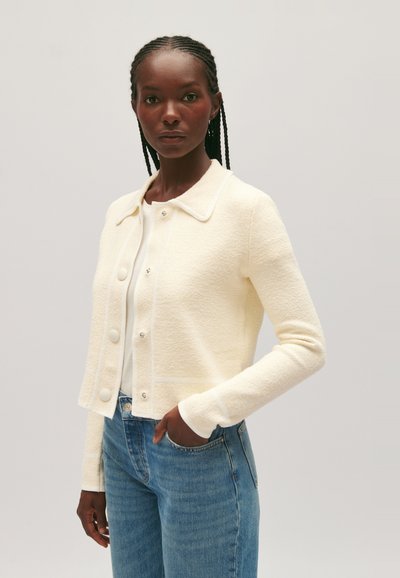 Mujer con el cabello trenzado, llevando una chaqueta texturizada color crema con botones sobre una blusa blanca y jeans azules, de pie contra un fondo plano.