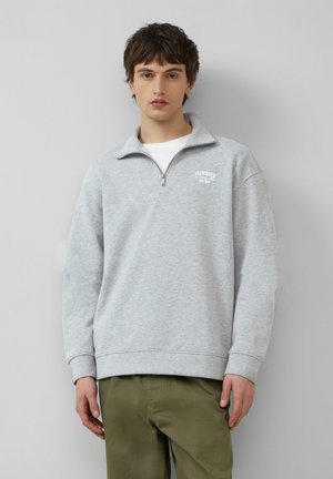 Helle graue Pullover-Sweatshirt mit einem Viertelreißverschluss, gerippten Bündchen und Saum. Mit einem kleinen weißen Logo auf der Brust. Über einem weißen T-Shirt getragen.