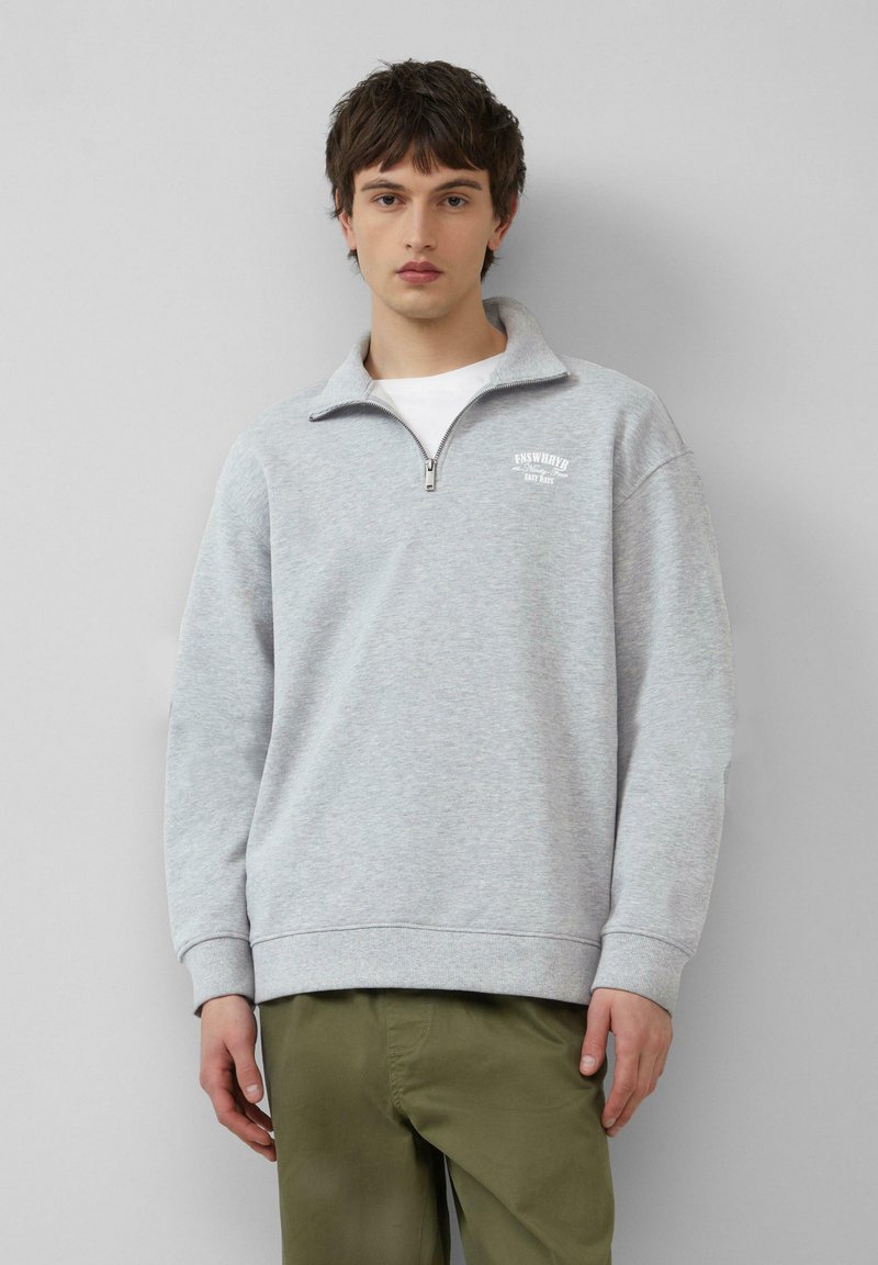 Felpa leggera grigia con zip a quarto, polsini e fondo a costine. Presenta un piccolo logo bianco sul petto. Indossata sopra una maglietta bianca.