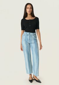 Top ajustado negro de manga corta con botones, combinado con jeans de pierna ancha de color azul claro que presentan un patrón de rayas onduladas. Zapatos planos negros con punta.