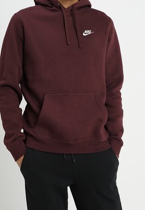 Bordowa bluza Nike z kapturem na troczki i przednią kieszenią, noszona z czarnymi spodniami, ręce częściowo w kieszeniach.