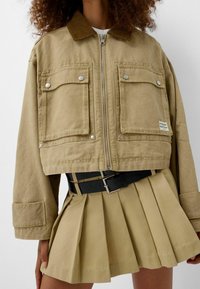 Skrócona beżowa kurtka z sztruksowym kołnierzem, dwoma kieszeniami na piersi, srebrnymi detalami i teksturowanym materiałem. Nosi się ją na plisowanej beżowej spódnicy.