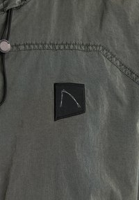 Chaqueta de tela verde, ligera, con una superficie texturizada. Presenta un bolsillo de parche negro y un diseño bordado. Incluye capucha con cordón.