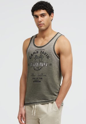 Mann mit lockigem schwarzen Haar, der ein grünes ärmelloses Tanktop mit schwarzem Schriftzug und beiger Hose trägt, steht mit einer Hand in der Tasche.
