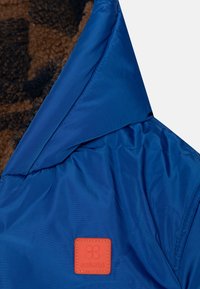 Niebieska pikowana kurtka z brązowym futerkiem od wewnątrz. Posiada wyprofilowany kołnierz i pomarańczową naszywkę z logo. Gładki materiał zewnętrzny.