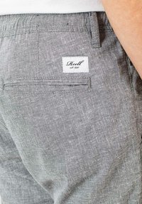 Gray linen shorts with a textured finish, featuring a back pocket with a white label reading "Reell est. 1997" sewn onto it.