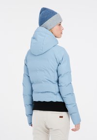 Lichtblauwe pufferjas met een getailleerd silhouet, capuchon en zwart ribgebreid zoom; gedragen met een blauw en grijs gebreid muts.