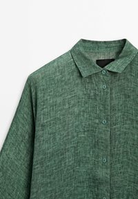 Chemise en lin verte avec col pointu, fermeture à boutons, et tissu texturé avec un motif tissé subtil.