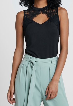 Donna con top nero senza maniche con dettagli in pizzo e scollo a goccia, abbinato a pantaloni verde chiaro a vita alta con cintura annodata.