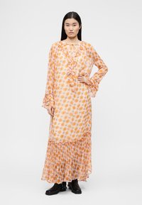 Γυναίκα που στέκεται, φοράει μακρύ, διαφανές, φλοράλ πορτοκαλί και ροζ φόρεμα με βολάν, συνδυασμένο με μαύρα μποτάκια αστραγάλου σε απλό φόντο.