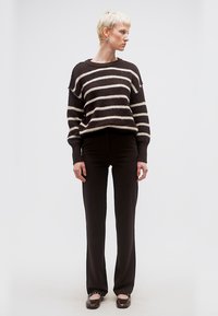 Γυναίκα με κοντά ξανθά μαλλιά που φοράει ασπρόμαυρο ριγέ πουλόβερ, μαύρο παντελόνι και μαύρα παπούτσια, όρθια και κοιτάζει προς τα πλάγια.