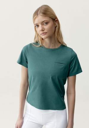 Mladá žena s dlouhými blond vlasy, oblečená do tyrkysově modrého trička s krátkými rukávy a malou náprsní kapsou a bílých kalhot, na jednobarevném pozadí.