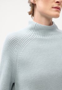 Svjetloplava, teksturirana pletena vestu s visokim ovratnikom i raglan rukavima. Prikaz izbliza usredotočen na detalje tkanine i ovratnika.