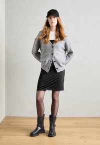 Cardigan en tricot gris avec des boutons, robe noire ajustée, collants noirs transparents, bottines noires avec détail de boucle, et casquette noire avec logo.