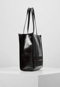 Lśniąca czarna prostokątna torba na ramię z długimi uchwytami i wytłoczonym napisem "Versace Jeans Couture" na przodzie, wyeksponowana na białym piedestale.