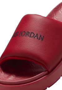 Sandale rouge à glissière avec un dessus lisse semblable à du cuir. Présente un texte en relief « AIR JORDAN » en noir et un design de semelle texturé.