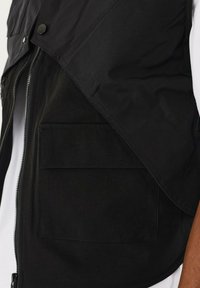 Sort vest med lynlåsåbning, stor klaplomme og struktureret stof. Designet med et lagdelt udseende og glat finish.