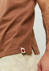 Hnedé bavlnené tričko s bočnými rozparkom, s malou červenou bodkou na leme. Jemná textúra, priliehavý strih.