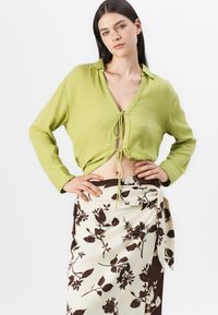 Chemise verte lime texturée avec un col en V et une fermeture à nouer à la taille, accompagnée d'une jupe portefeuille à motifs floraux crème et brun.