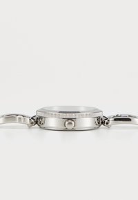 Stainless steel wristwatch case with ridged bezel and crown, connected to metal watchband links on both sides, shown from side view.