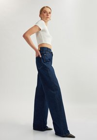 Top blanco de algodón recortado combinado con pantalones de mezclilla de pierna ancha de cintura alta en azul oscuro, que cuentan con cinco bolsillos y delicados detalles de costura.