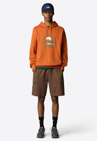 Felpa arancione con cappuccio e stampa grafica, pantaloni cargo marroni, calze scure e scarpe da escursione nere. Il cappellino è blu navy con logo.