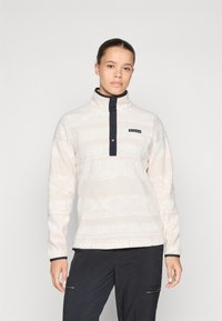 Fleece pullover in lichtbeige met subtiele witte patronen, hoge kraag en marineblauwe accenten. Voorzien van een knoopsluiting aan de voorkant en logo detail.