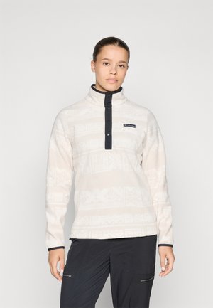 Fleece genser i lys beige med subtile hvite mønstre, høy krage og marineblå detaljer. Har frontklikk og logo detaljer.