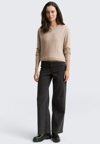 Vrouw staat in een beige lange mouwen trui, zwarte wijde jeans en zwarte stevige laarzen tegen een effen lichte achtergrond.
